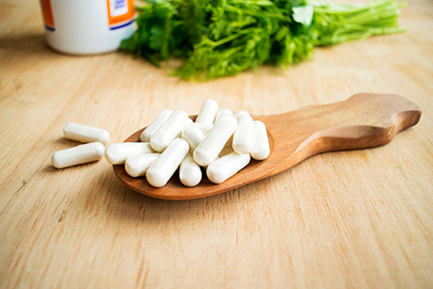 Health effects of coagulations vitamin. White capsules of vitamin K (phylloquinone) in wooden spoon on a background of a bottle of tablets and green herbs. Vitamins supplements in spoon on wooden table