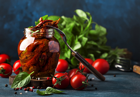 Italian Sun Dried tomatoes in olive oil with green basil and spices in glass jar on blue kitchen table, copy space