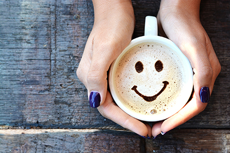 Smiley face on cappuccino foam, woman hands holding one cappuccino cup on wooden table